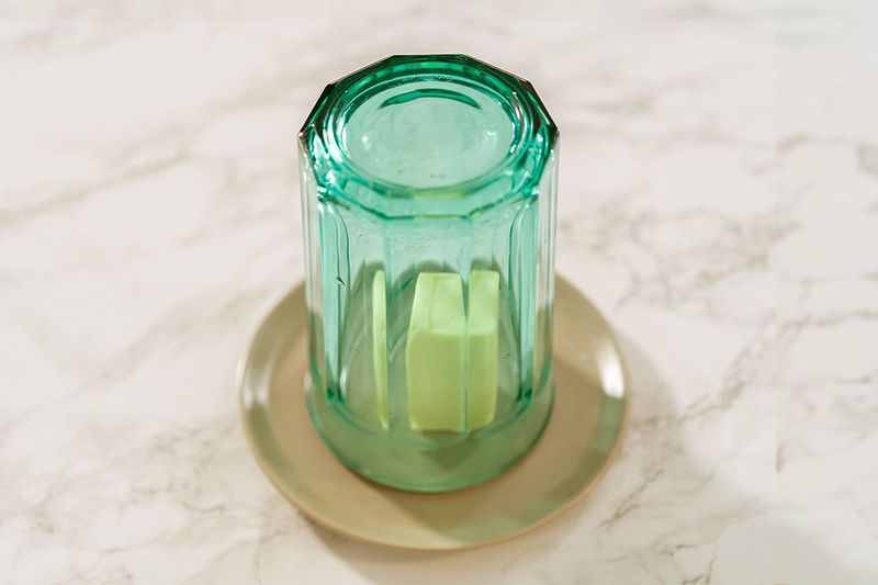A green drinking glass placed upside down to cover a pat of butter that's sitting on a small, beige plate on a marble countertop.