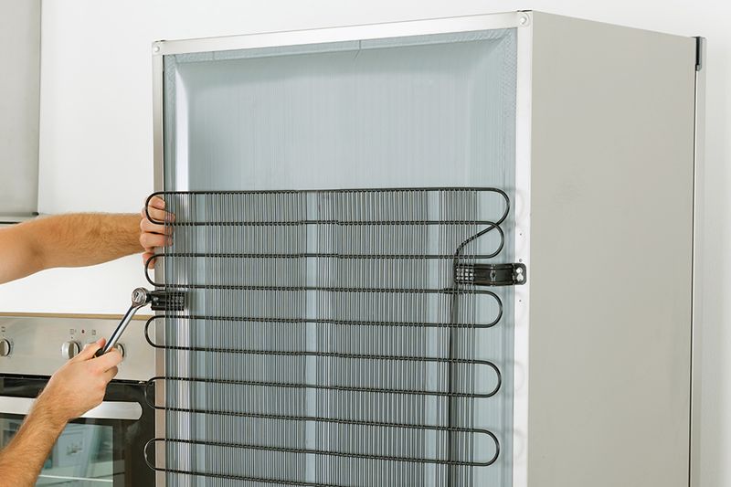 An anonymous person tightening a part on the back of a refrigerator that keeps the coils attached. The refrigerator has very clean coils and is in a kitchen.