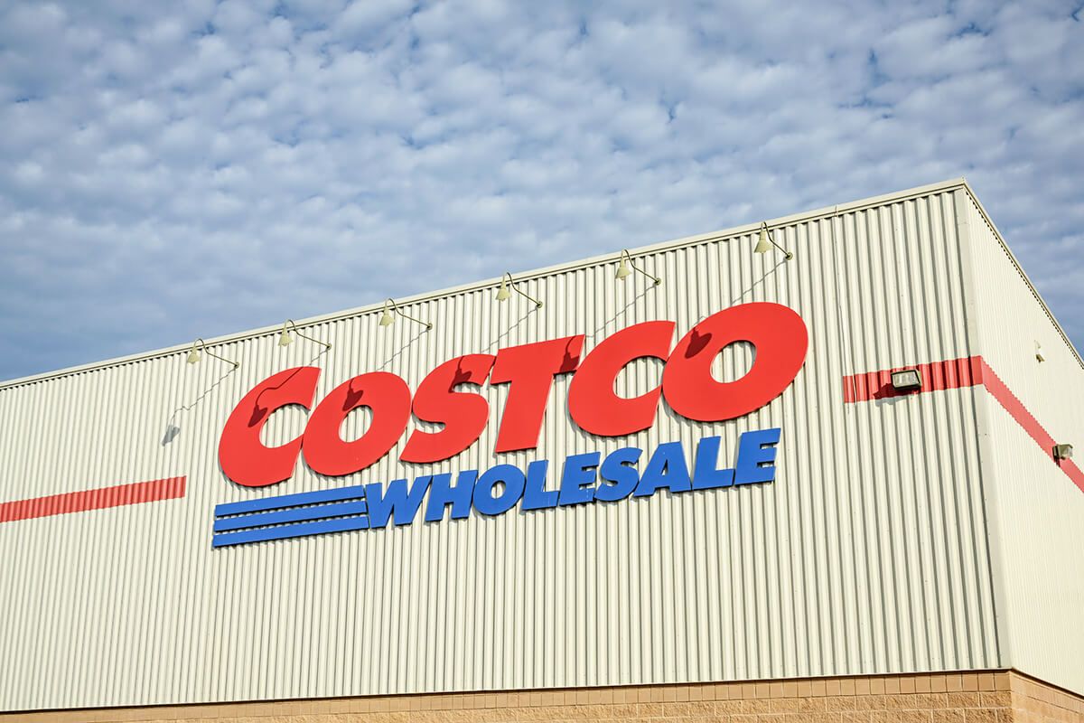 Close-up of the Costco Wholesale logo on the exterior of one of their warehouses. The photo was taken during the daytime and there are lots of clouds in the sky.