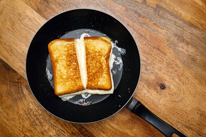 A perfect grilled cheese made with white cheddar in a black nonstick frying pan on a wooden countertop.