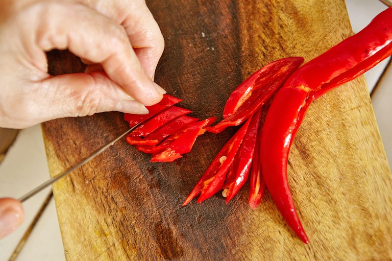 Close-up overhead shot of someone slicing red chili peppers on a wooden cutting board.