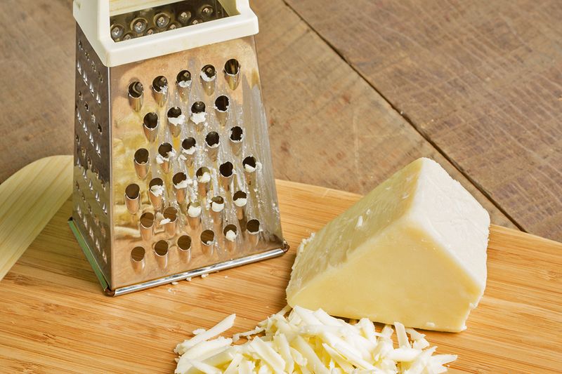 A block of white cheese on a wooden cutting board with some shavings next to it and a box grater.