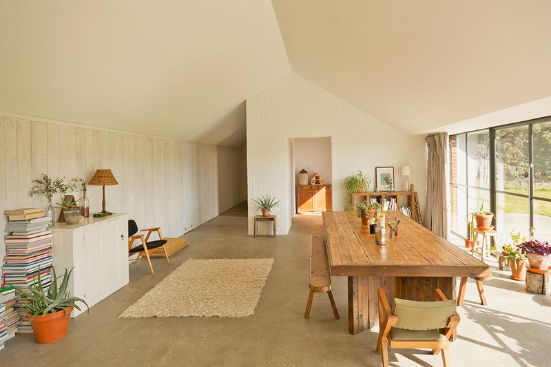 Open-concept dining room with tall ceilings. There's a large wooden dining table, a small shag rug, a white cabinet, stacks of books, and plants.