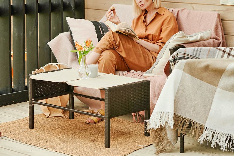 Anonymous woman eating a snack and reading a book on a small patio with a loveseat, coffee table, and an accent chair.