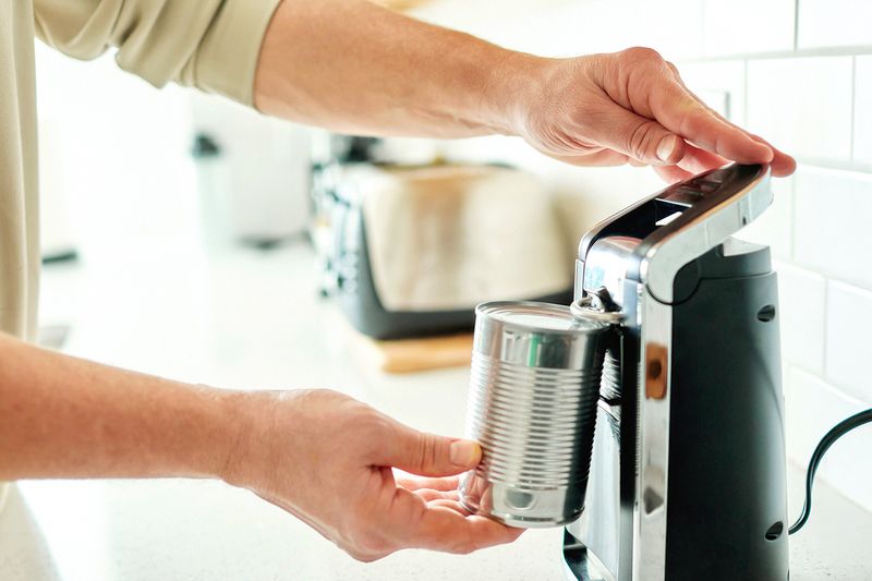 An anonymous person (showing the arms/hands only) using an automatic can opener in a kitchen.