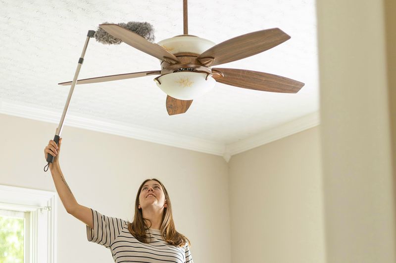 Woman using an extendable duster to clean the blades of a ceiling fan.