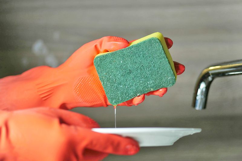 Close-up of someone wearing orange cleaning gloves holding a sponge dripping with water over a small plate with the faucet in the background.