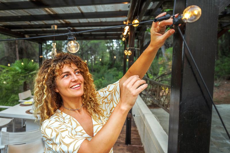 A blonde woman with curly hair adjusting string lights on an outdoor pergola.