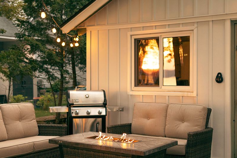 A backyard patio at dusk with string lights, two loveseats, a fire pit, and a barbecue.
