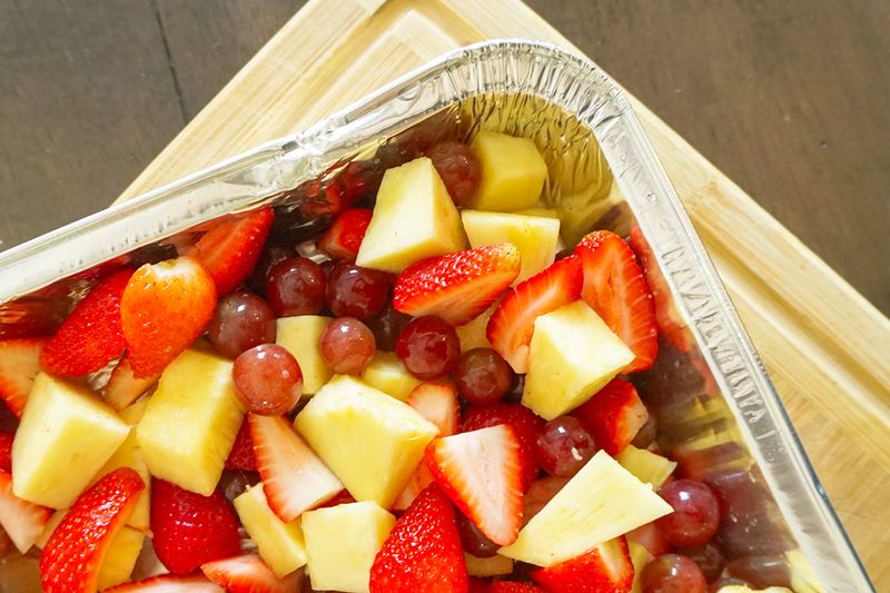 A close-up of a disposable aluminum tray filled with cut fruit, including strawberries, pineapple, and red grapes.