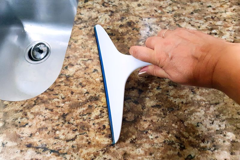 Anonymous person using a squeegee to clean granite kitchen countertops, pushing the excess water into a nearby sink.
