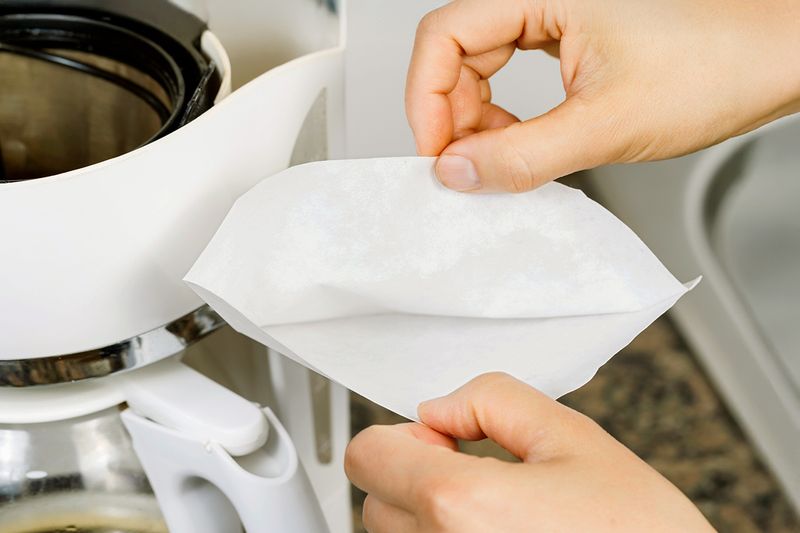 A close-up of a person's hands opening up a white, cone-shaped coffee filter next to a drip coffee maker.