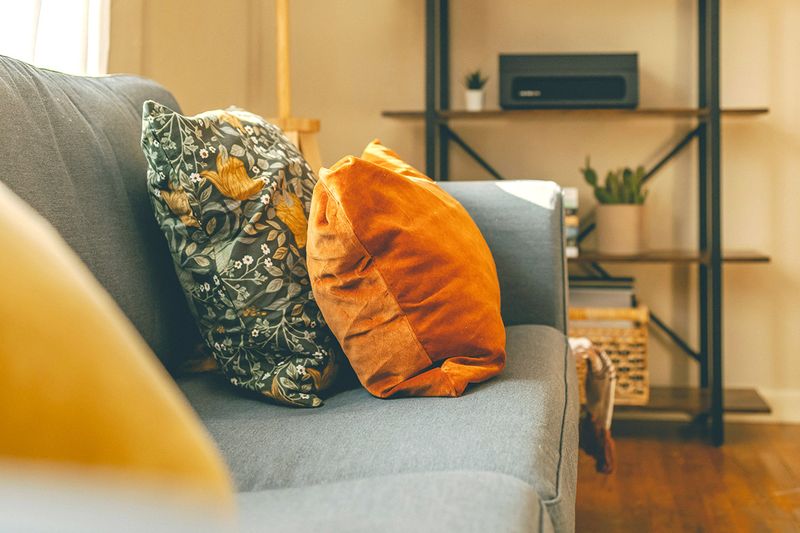 Two in-focus throw pillows on a gray couch. One is orange velvet, and the other is teal with a botanical print.