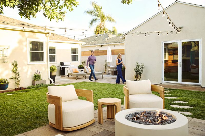 A well-designed backyard with a paved patio area featuring loungers, a grill, and dining table. Plus, a grassy area with a stone path, plush chairs, and a modern fire pit.
