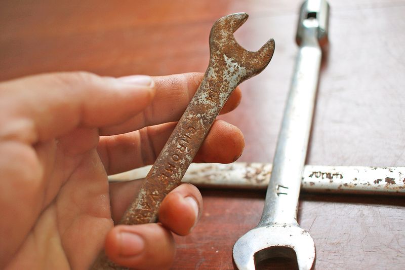 Anonymous person's hand holding a small rusty wrench. In the background is a similar sized wrench that has been cleaned, with just a few rust spots remaining.