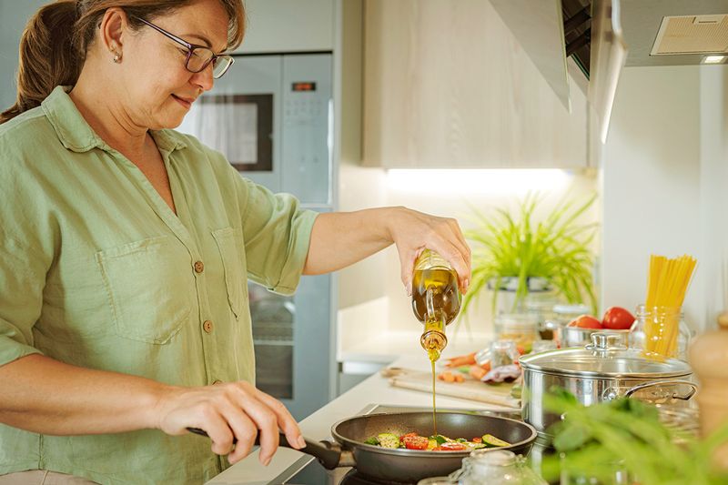 Woman pouring cooking oil into a frying pan with cut vegetables. The frying pan is on an electric cooktop and there are various ingredients in the background.