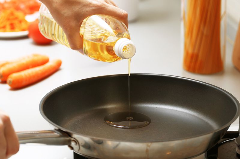 Anonymous person adding oil from a bottle to a frying pan. In the background are three whole carrots.