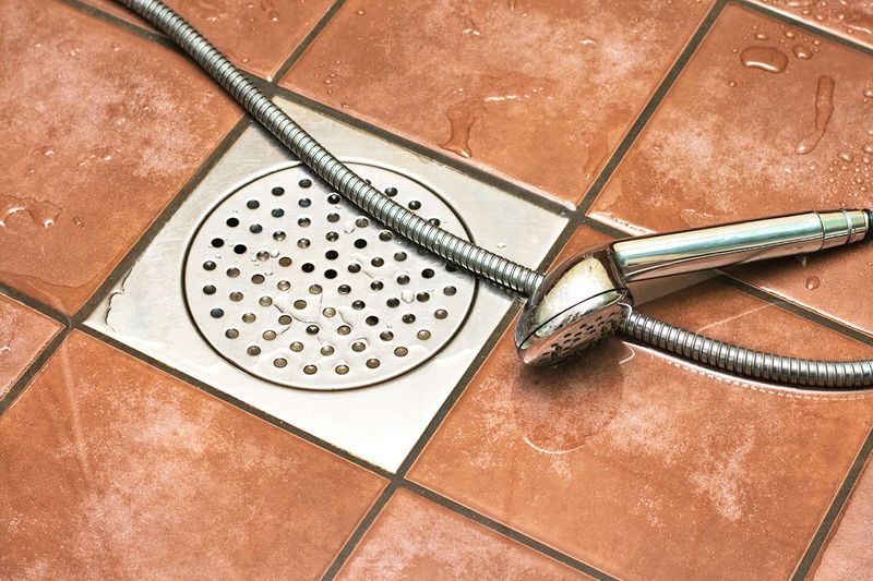 A shower floor with terracotta-colored square tiles and a stainless steel drain. On the floor is a handheld shower head.