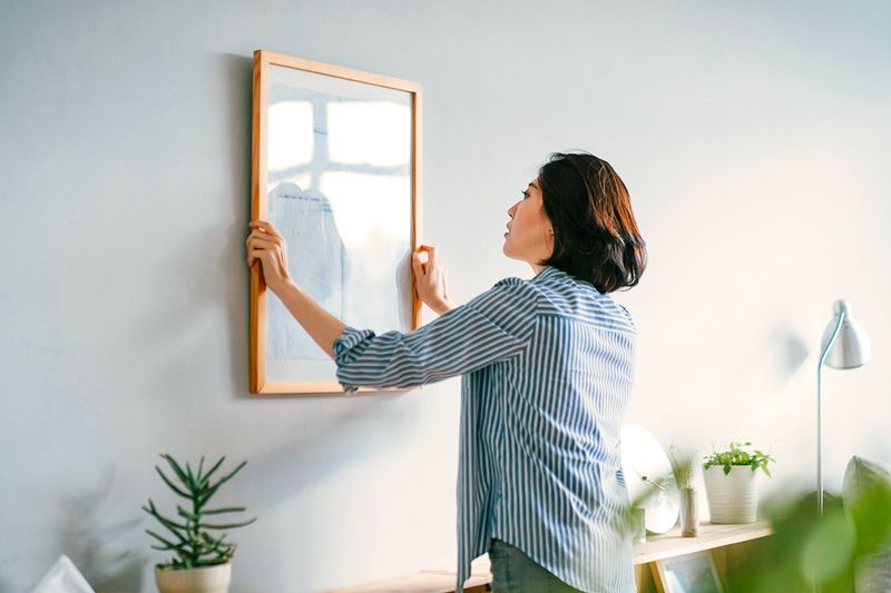 Woman hanging a framed piece of art on a blue wall.