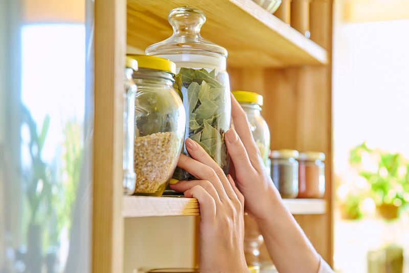 Anonymous person reaching for a jar of dried bay leaves in a clear glass jar in the pantry.