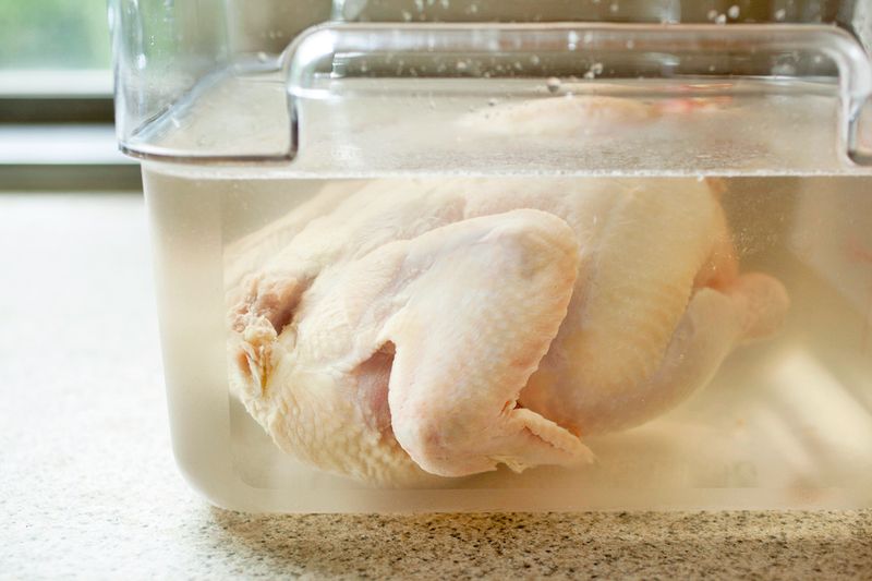 A whole, raw chicken brining in a clear container, viewed from the side.