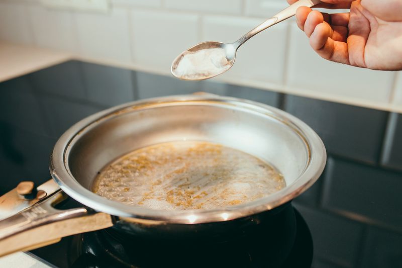 Anonymous person sprinkling baking soda into a stainless steel pan with a burnt bottom.