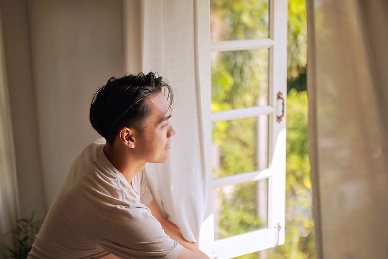 Young man looking out through an open window. The window is framed by tan linen curtains.