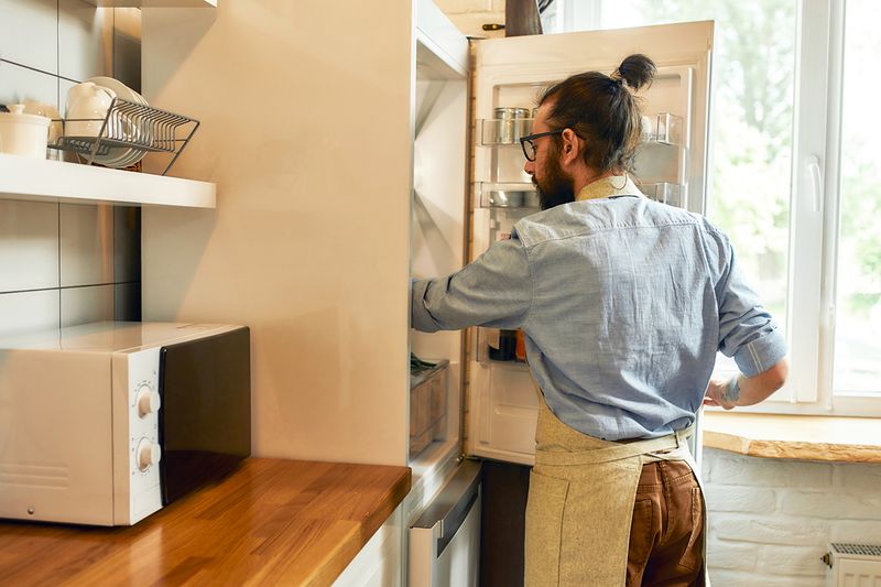 A young, bearded man wearing an apron reaching into a refrigerator to organize the food inside.