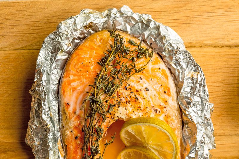 A filet of cooked salmon unwrapped from a foil packet. On the salmon are a few sprigs of thyme and two lemon slices.