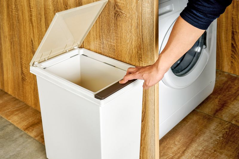 Anonymous person pushing the lever of a white trash bin that's next to a washing machine.