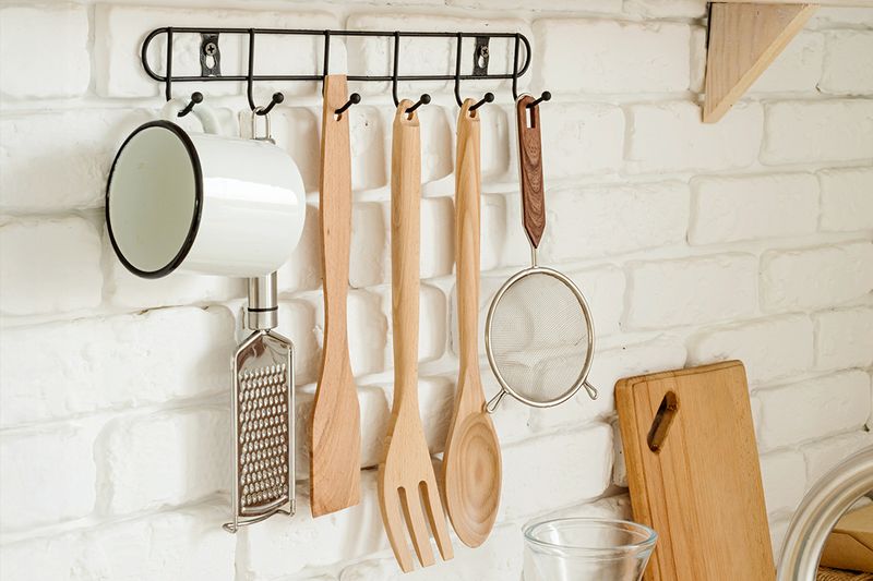 A close-up of kitchen utensils hanging from wall-mounted hooks. The wall is made of white brick.