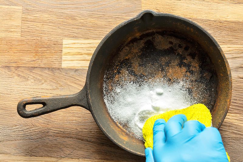 An anonymous person wearing cleaning gloves is scrubbing the rusty cooking surface of a cast iron pan with salt and a yellow scouring pad.