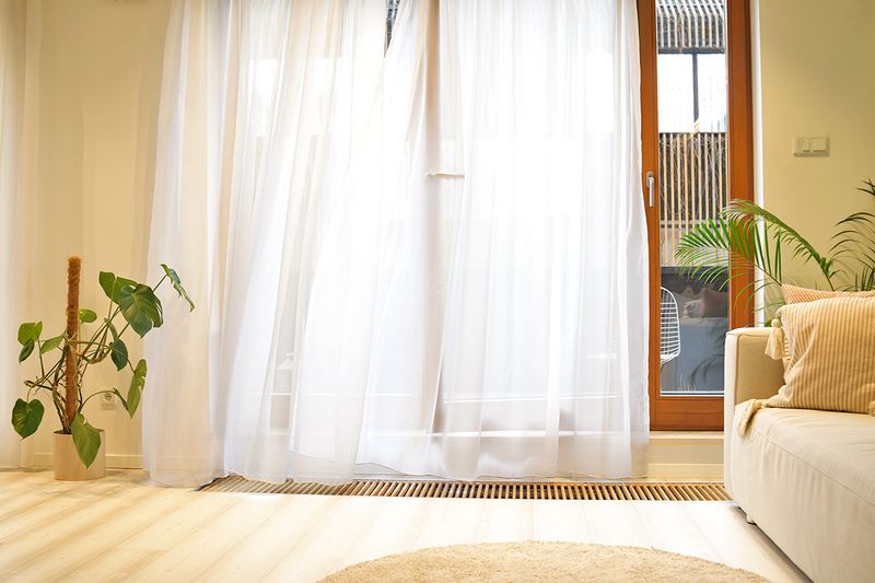 A cracked-open door to a patio behind white sheer curtains that are billowing in the wind.