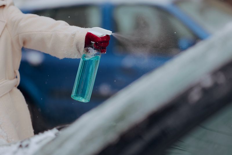 Anonymous person spraying a blue solution in a spray bottle onto a windshield. The person is bundled up in winter clothing.