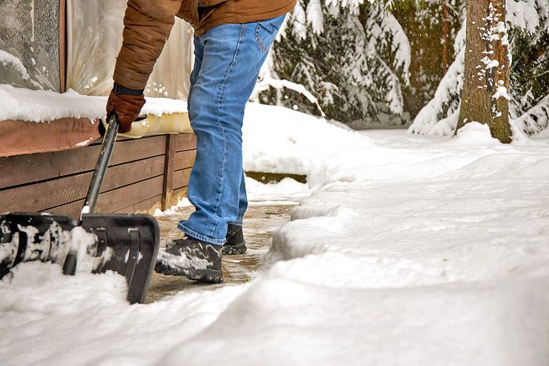 Anonymous person wearing jeans and a brown coat shoveling snow from a walkway in front of a house or business.