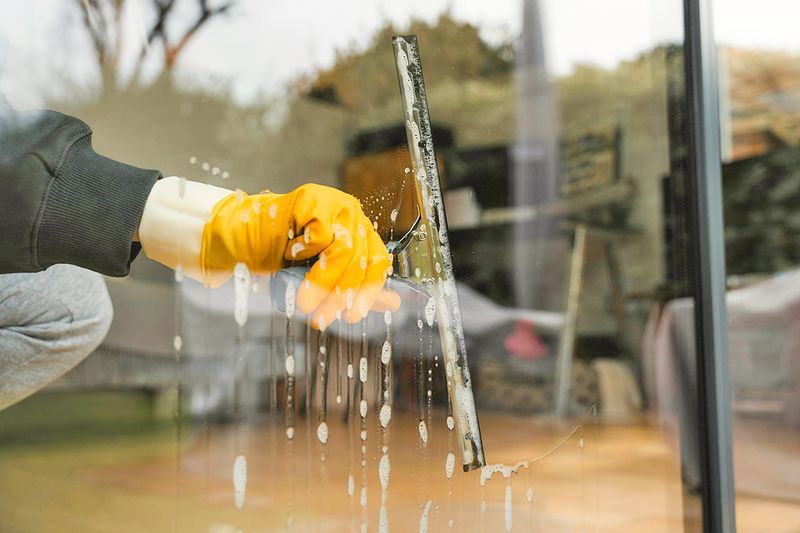 Anonymous person wearing yellow cleaning gloves washing a large window with a squeegee.