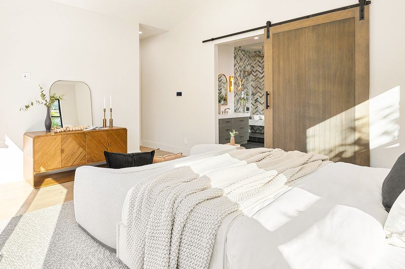 An all-white bedroom with a barn door leading to a bathroom with chevron pattern tile.