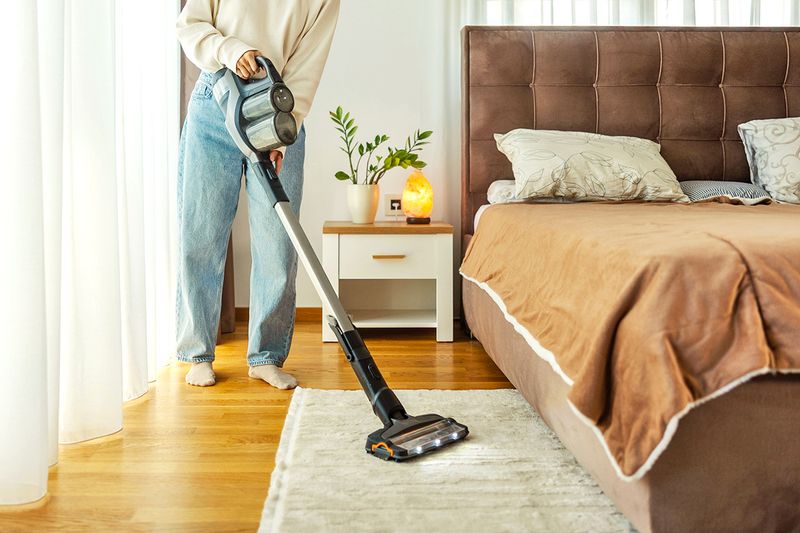 Anonymous woman vacuuming a rug underneath an upholstered bed.