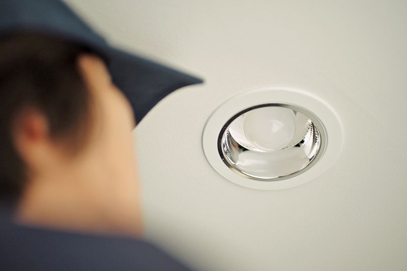 Anonymous person wearing a baseball hat looking up at a can light on the ceiling.
