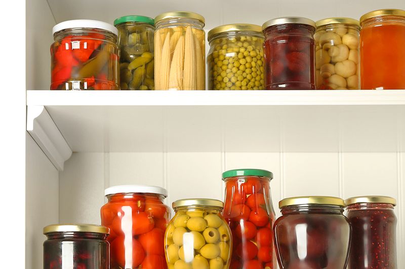 White-painted pantry shelves showing various canned ingredients, including olives, green beans, peppers, pickles, and peas.