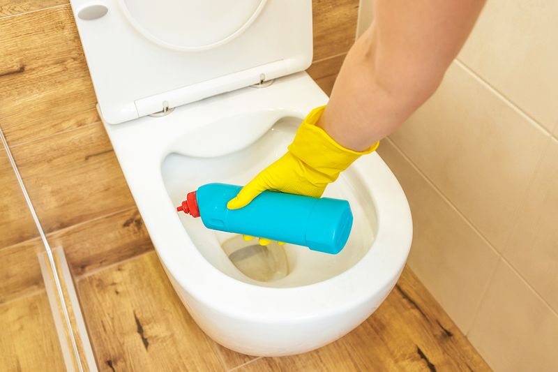 An anonymous person wearing cleaning gloves is applying a cleaning solution to a toilet bowl with yellow buildup rings.