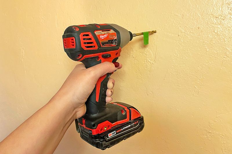 Anonymous person holding a red drill up to a beige wall. The drill bit has green painter's tape on it.