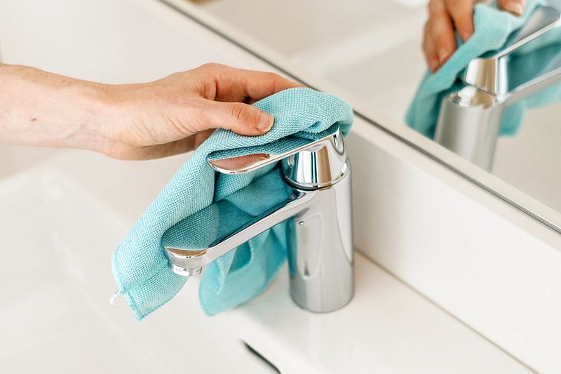 Anonymous person wiping a chrome bathroom faucet with a blue microfiber cloth.