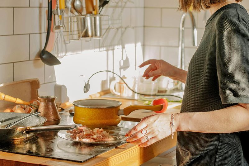 Anonymous woman following a recipe at her stovetop. In front of her is a saucepan, a skillet, and a plate of chopped, uncooked bacon.