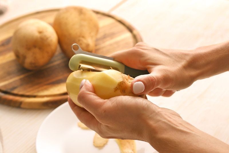 Anonymous person using a swivel peeler to peel a potato. At the top of the peeler is a potato eye remover tool.