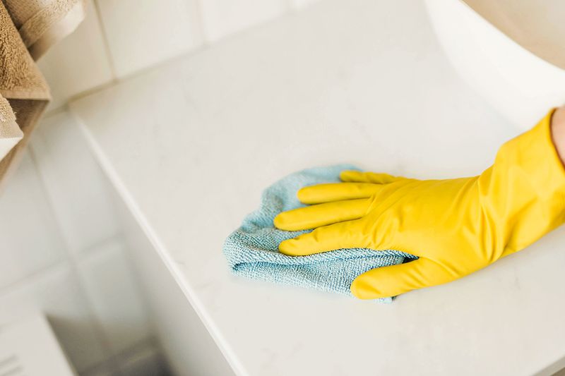 Anonymous person wearing a yellow cleaning glove wiping a countertop with a light blue microfiber cloth.