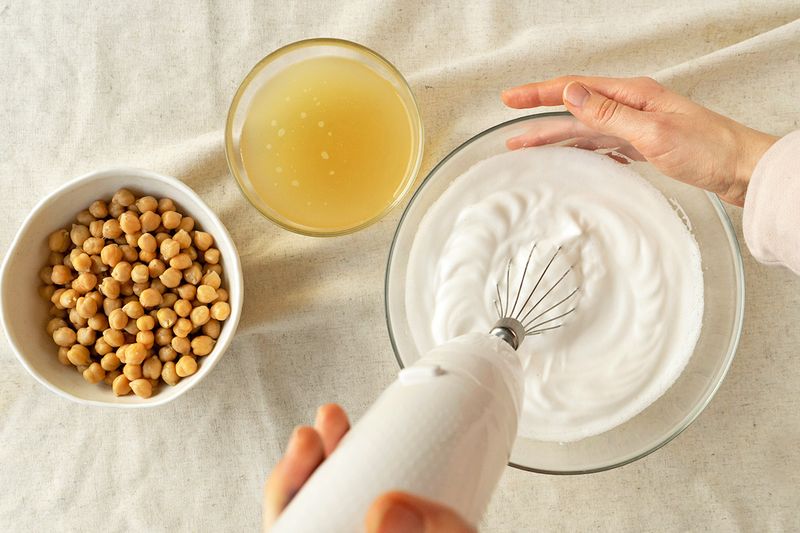 A bowl of chickpeas next to a bowl of aquafaba. An anonymous person is using an electric whisk to make eggless meringue.