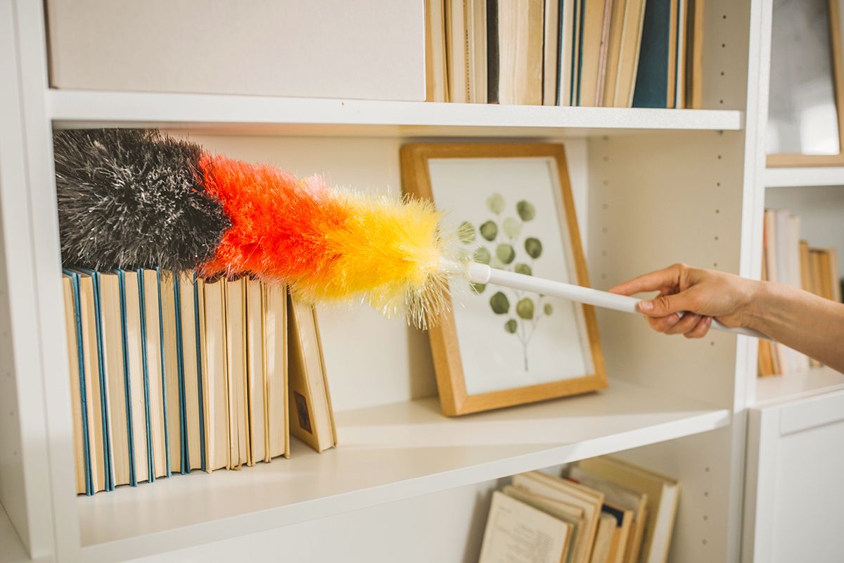 Anonymous person dusting a bookshelf with a black, red, and yellow duster.