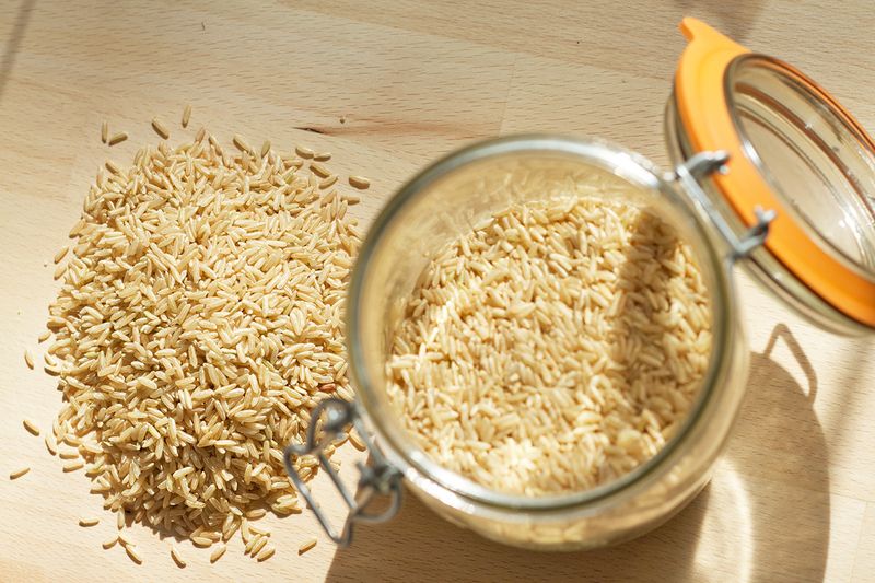 A glass canister of brown rice with some of the rice poured out onto the wood countertop beside it.
