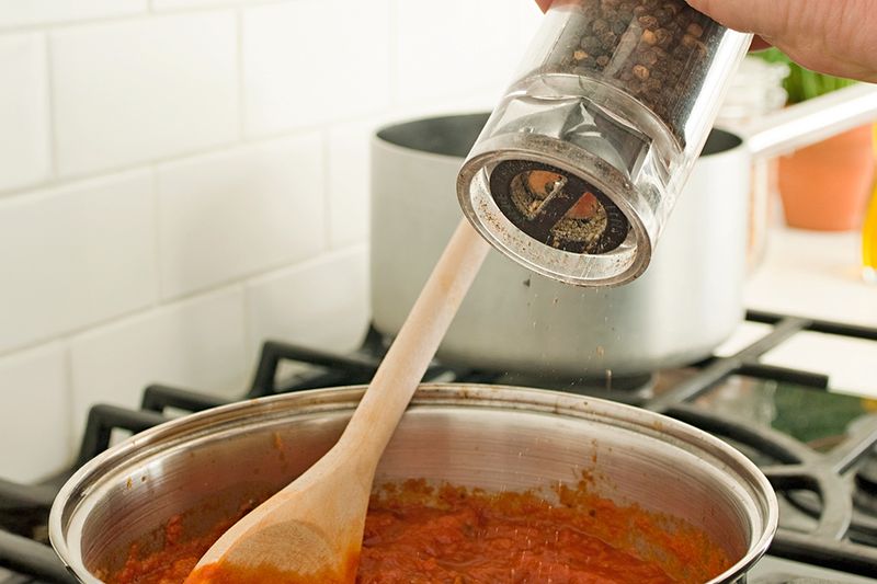 Anonymous person grinding pepper from a mill into a pot of red sauce on a stovetop.